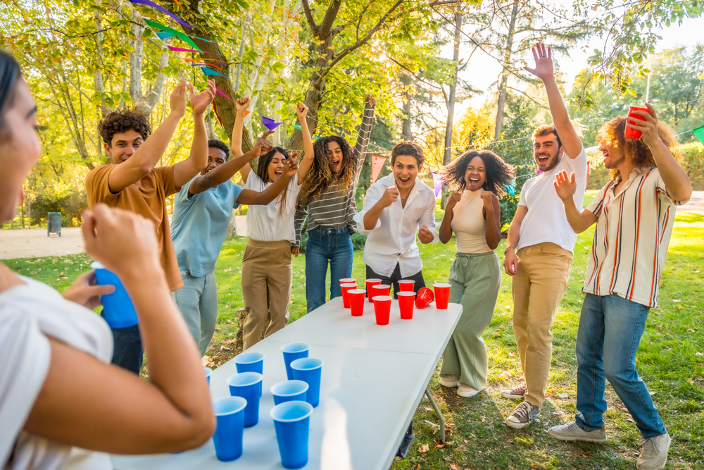 Young adults enjoying a lively game of beer pong while celebrating at a sunny park party, filled with laughter and camaraderie