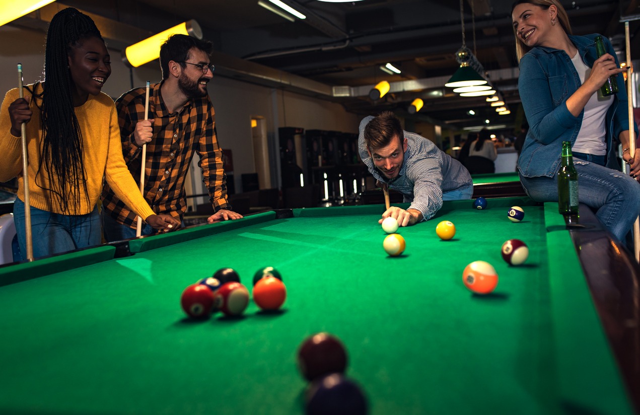 A group playng at a pool table, one taking a shot, two leaning on the table, one sitting on the edge of it, with a bottle on the table rim